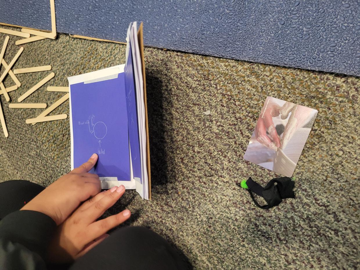 Hands point to the blue page of a small journal that has a What Is Learning card next to it.