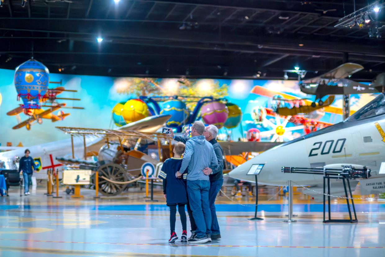 A small group stands in a large exhibit hall in an aerospace museum, there are large planes, rides, and simulators in the background