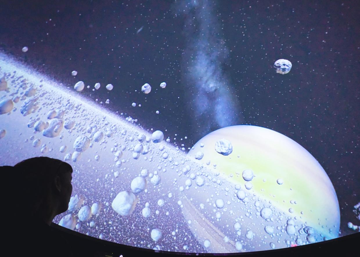 A shadow of a head gazes up at a bright planetarium screen, which is zooming in on an astroid belt.