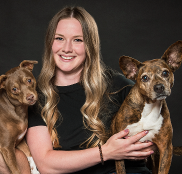 Woman in all black with long hair smiles with two adorable dogs, each brownish and sitting on either side
