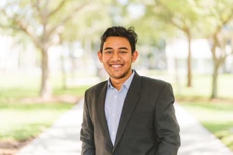 Headshot for Amdad Ahmed Awsaf; an individual stands among green trees on a sunny day. They are smiling, have short hair, and are dressed in business casual.