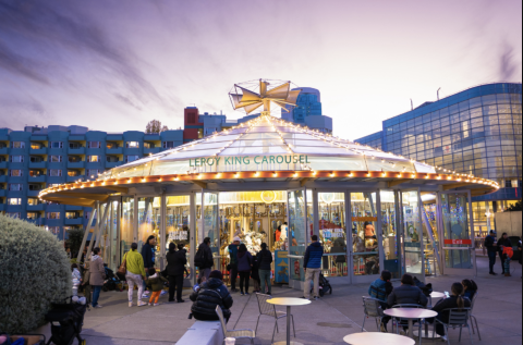 An image of a lighted carousel at night with people sitting in front at tables on a wide plaza