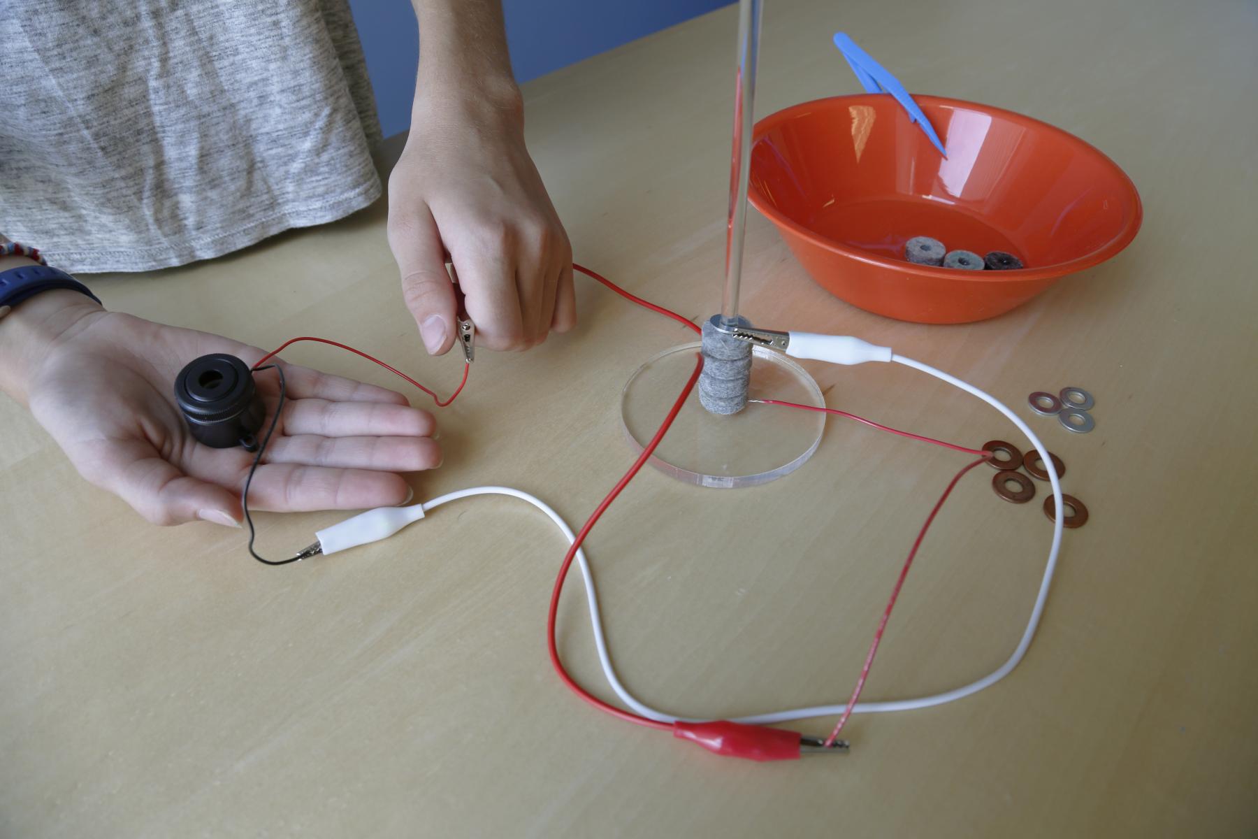 voltaic pile connected with wires to a buzzer held in someone's hand