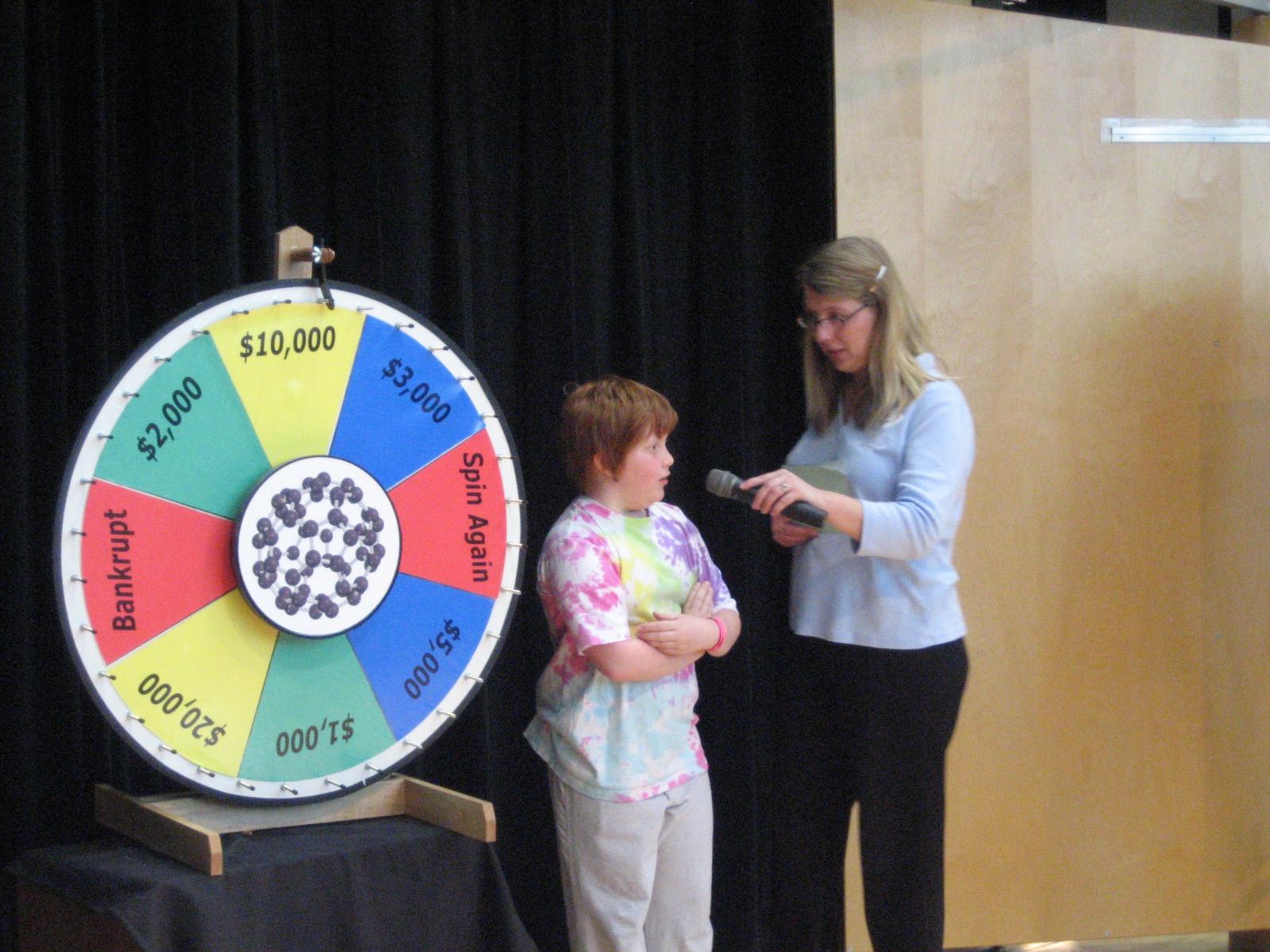 A facilitator on stage in front of a large spinning wheel asking a participant a question with a microphone
