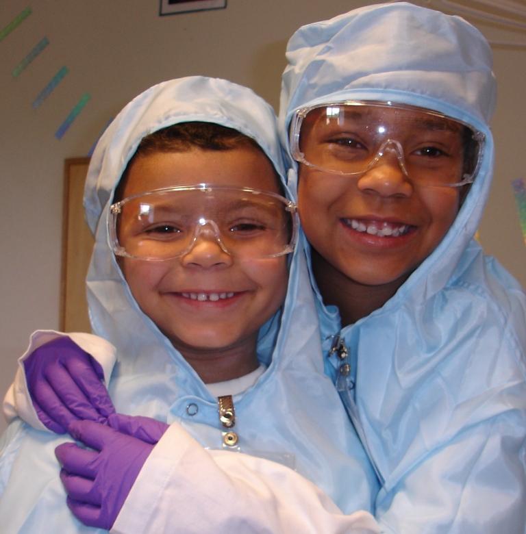 Two kids in cleanroom suits and PPE hugging and smiling at the camera