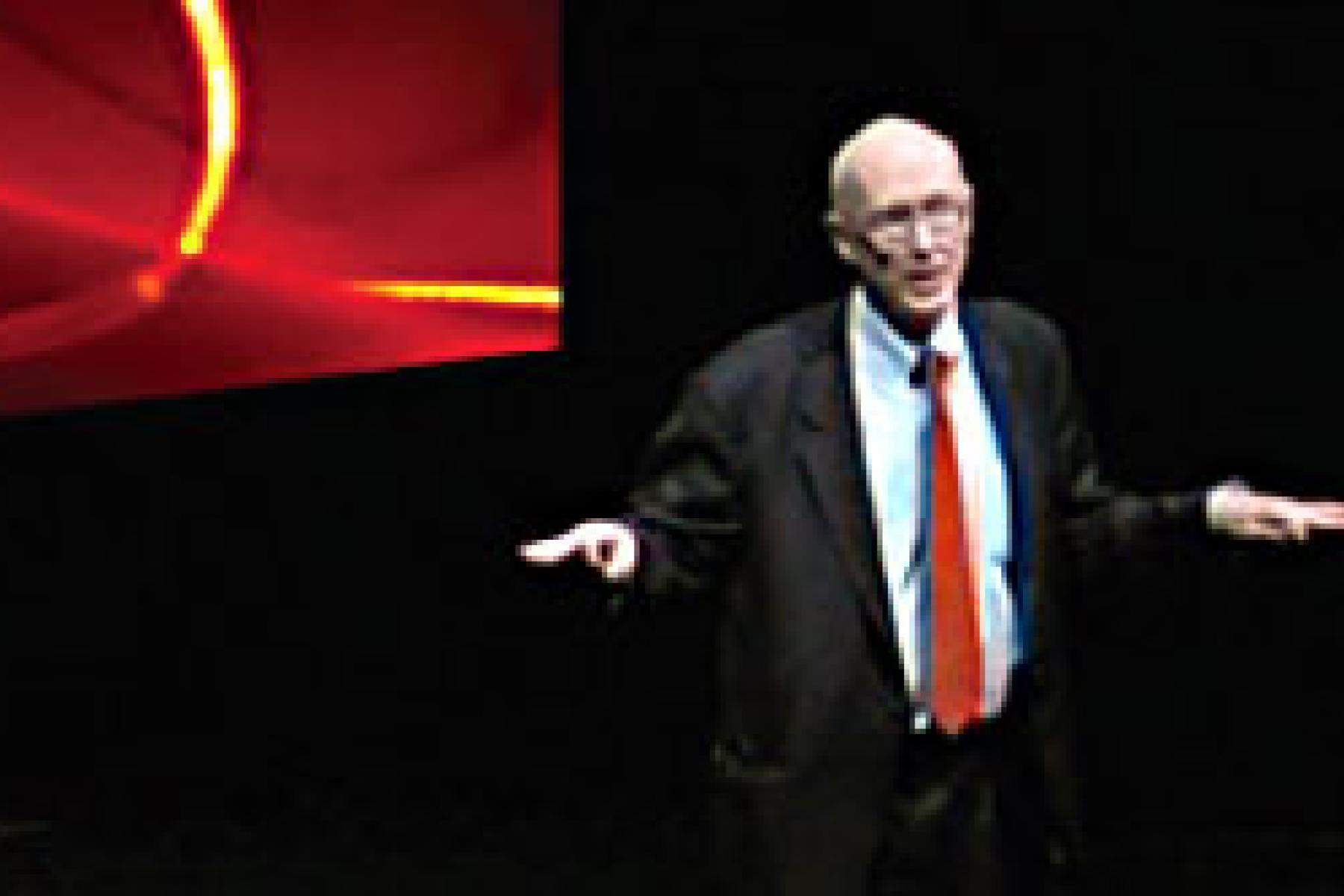 man with black coat, red tie and glasses speaks to an audience