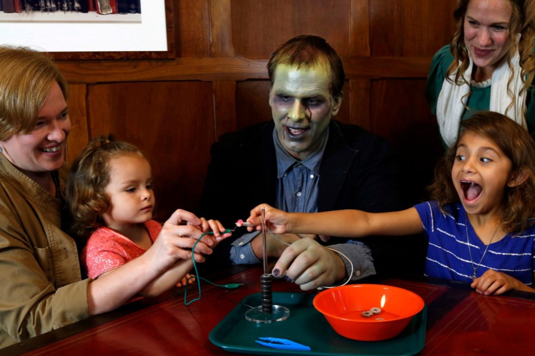 Two actors dressed as Mary Shelley and Frankenstein's Monster interact with a family of three demonstrating how to make a battery