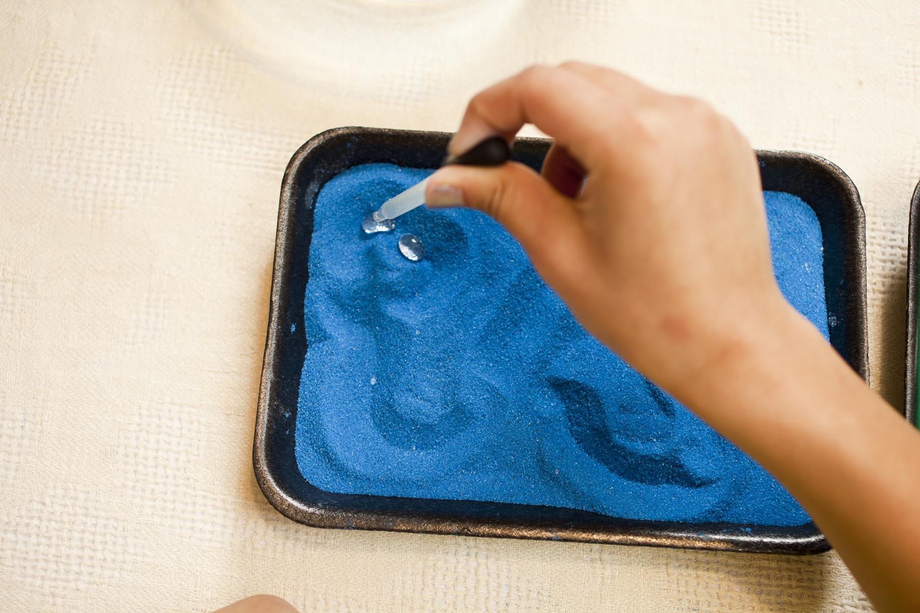 a person adds water droplets to blue colored sand using an eyedropper 