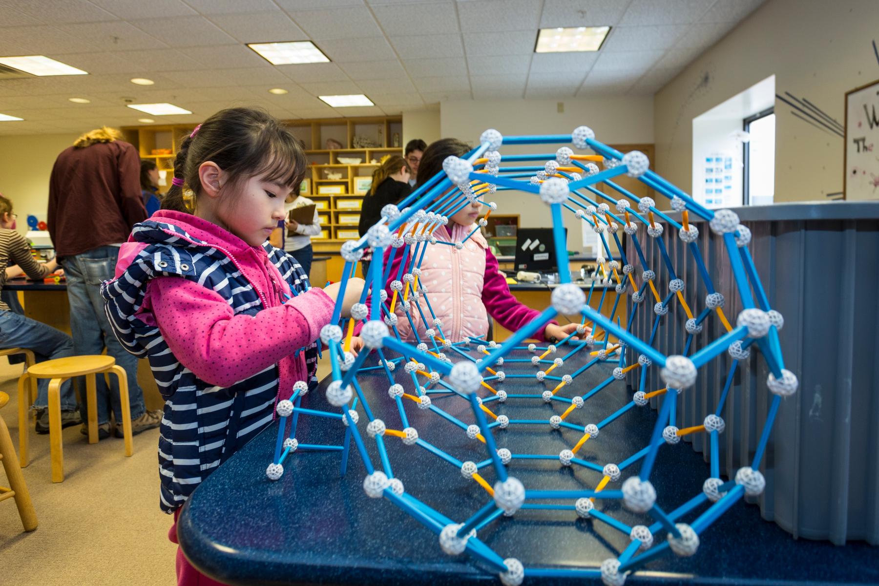 Young learner in a pink jacket assembles a nanotube model made of plastic rods and spheres