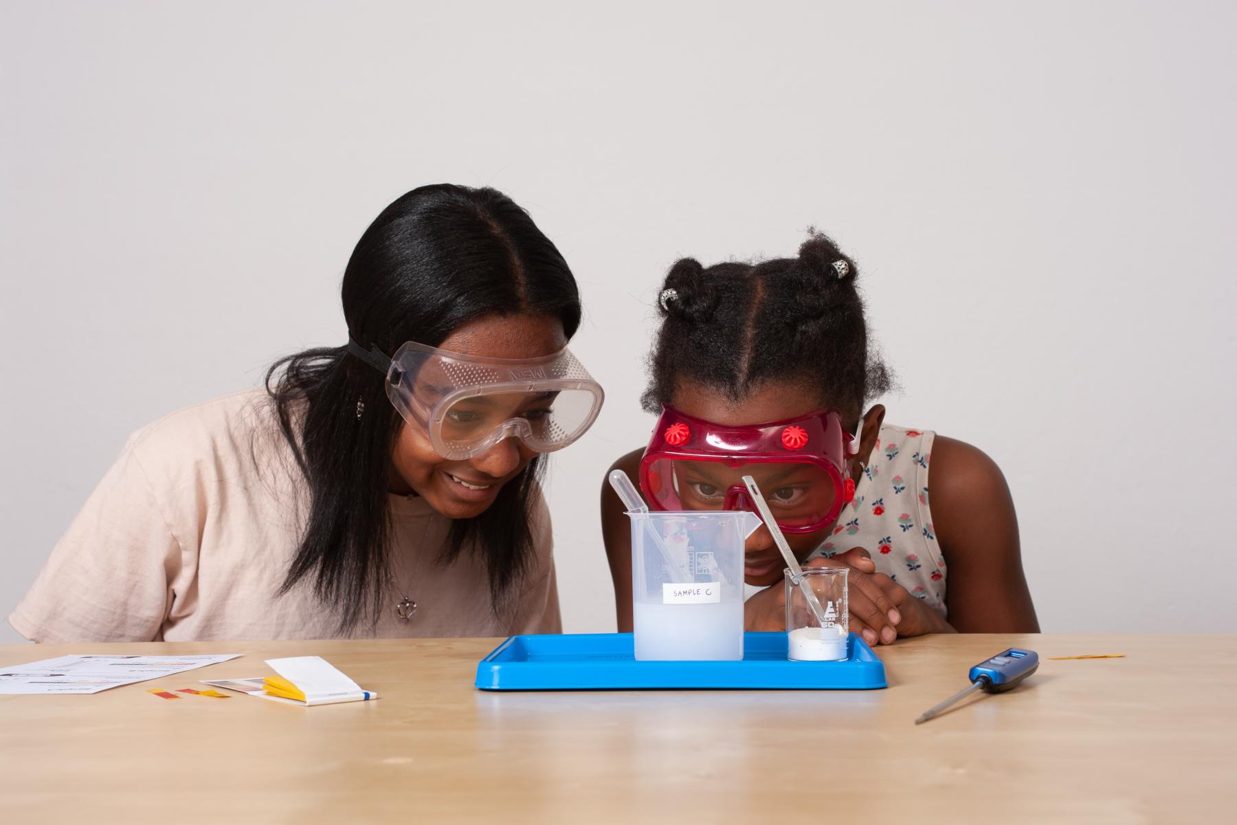 Two learners wearing goggles closely observe/read the measurement of the water line in a beaker