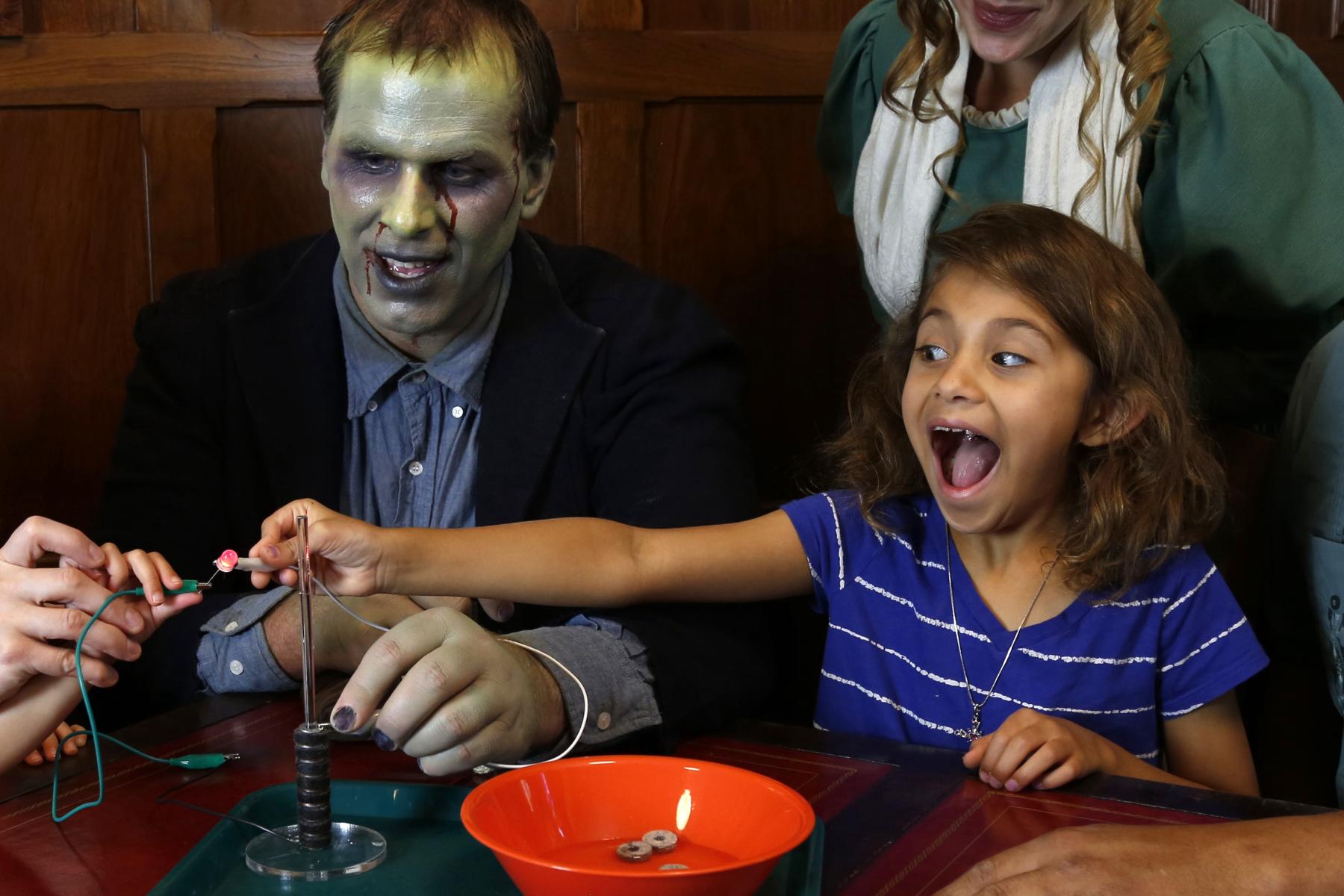 Child with excited facial expression using battery stack activity with adults dressed as Mary Shelley and Frankenstein's monster