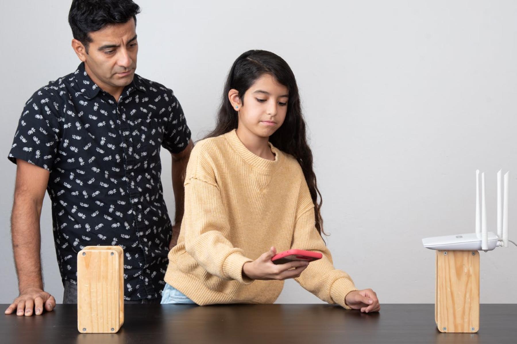 A child holds a smart phone in between two blocks of wood, placed apart on on a table, while an adult looks on. The block of wood on the right holds a Wi-Fi router. 