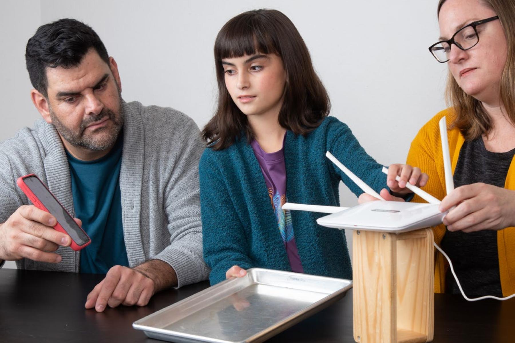 A child holds a metal baking sheet flat on a table in between two adults. The adult on the left holds a smartphone and the adult on the right is adjust the antennae of a Wi-Fi router on a block of wood.