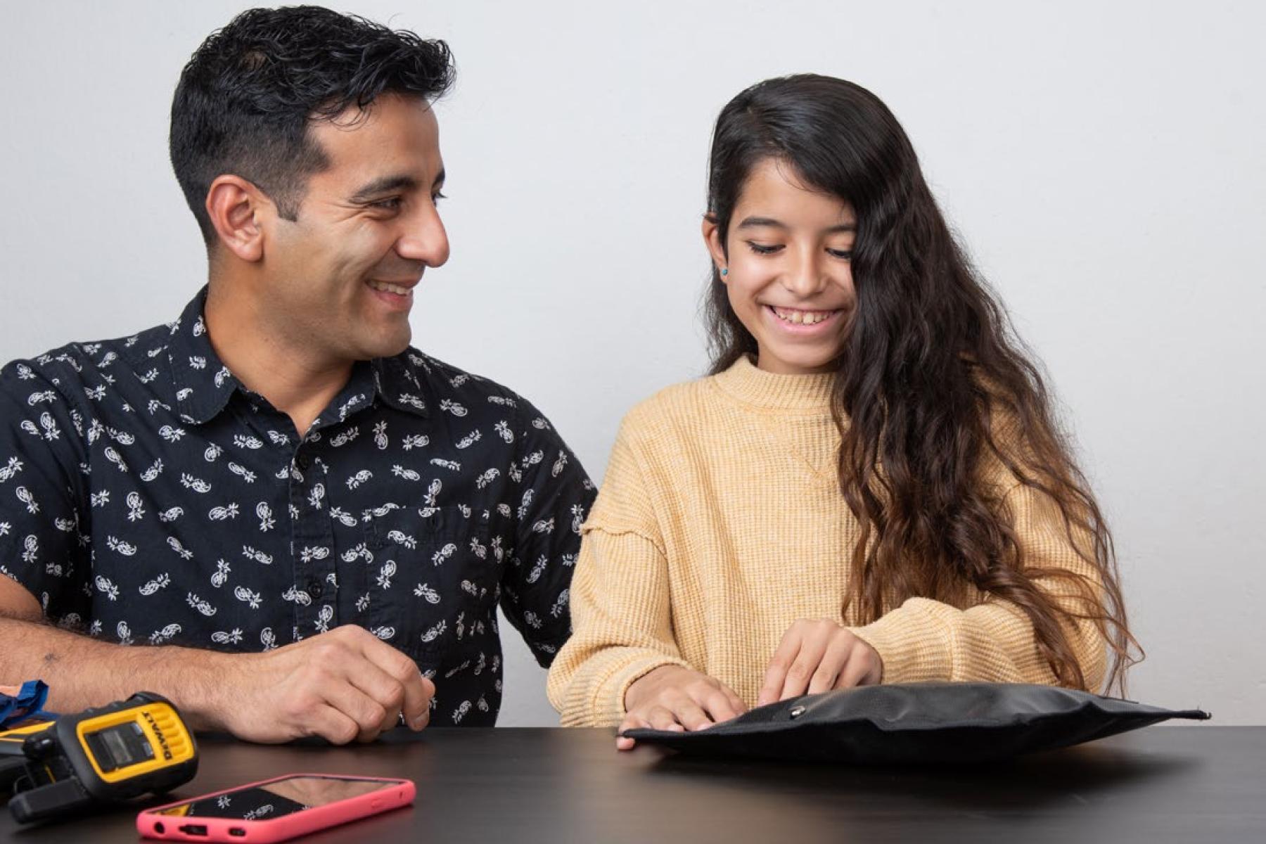 An adult and a child seated at a table both smile as the child wraps a radio device in a black bag called a Faraday bag. A walkie-talkie and a touch-screen mobile phone sit beside the bag.
