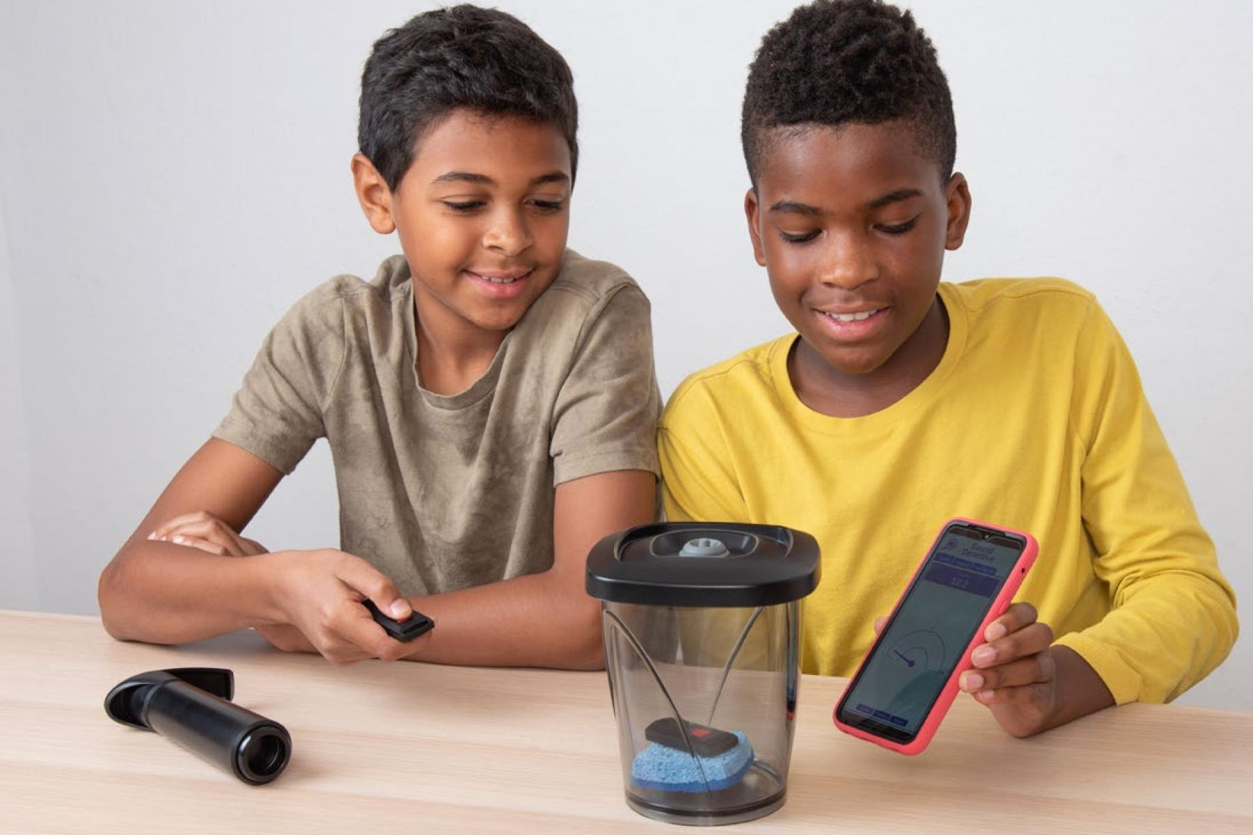 While sitting at a table, one child points a small transmitter at a vacuum-seal container that has a key finder inside, and another child holds a smartphone up to the other side of the container.