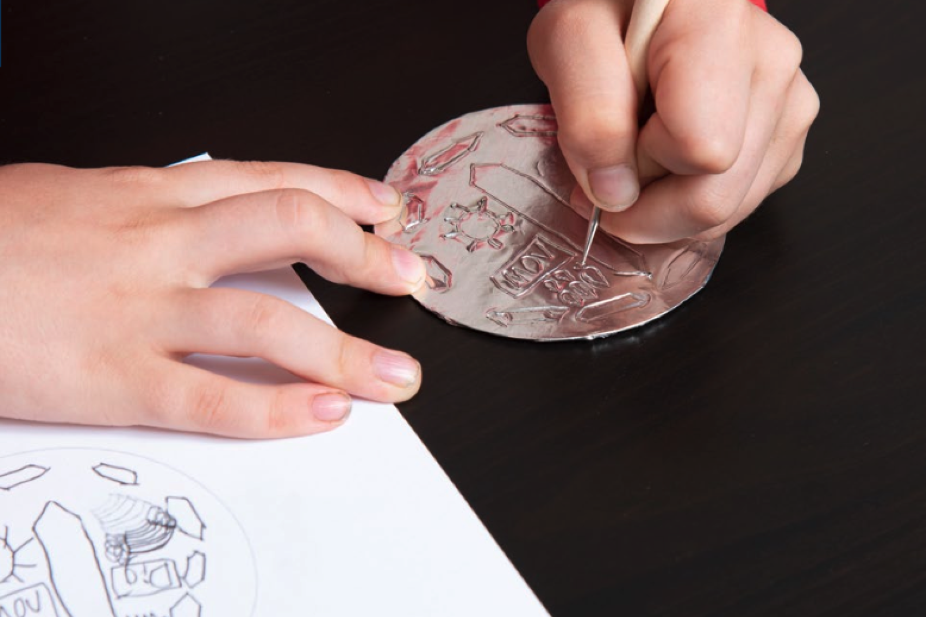 Hands Etch a Design into Foil as Part of the Moon Medallion Activity