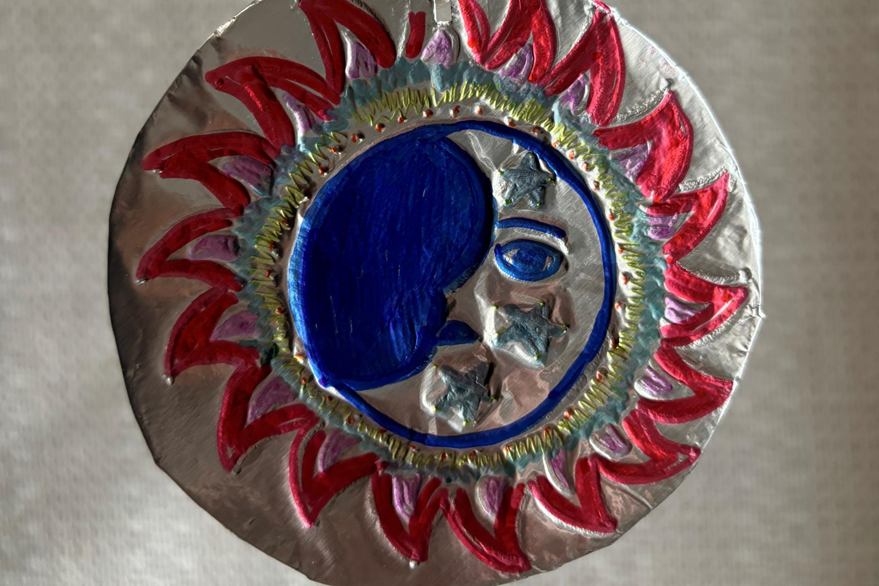 A colorful etching of the sun in tin foil with a string laced through the top in a loop.