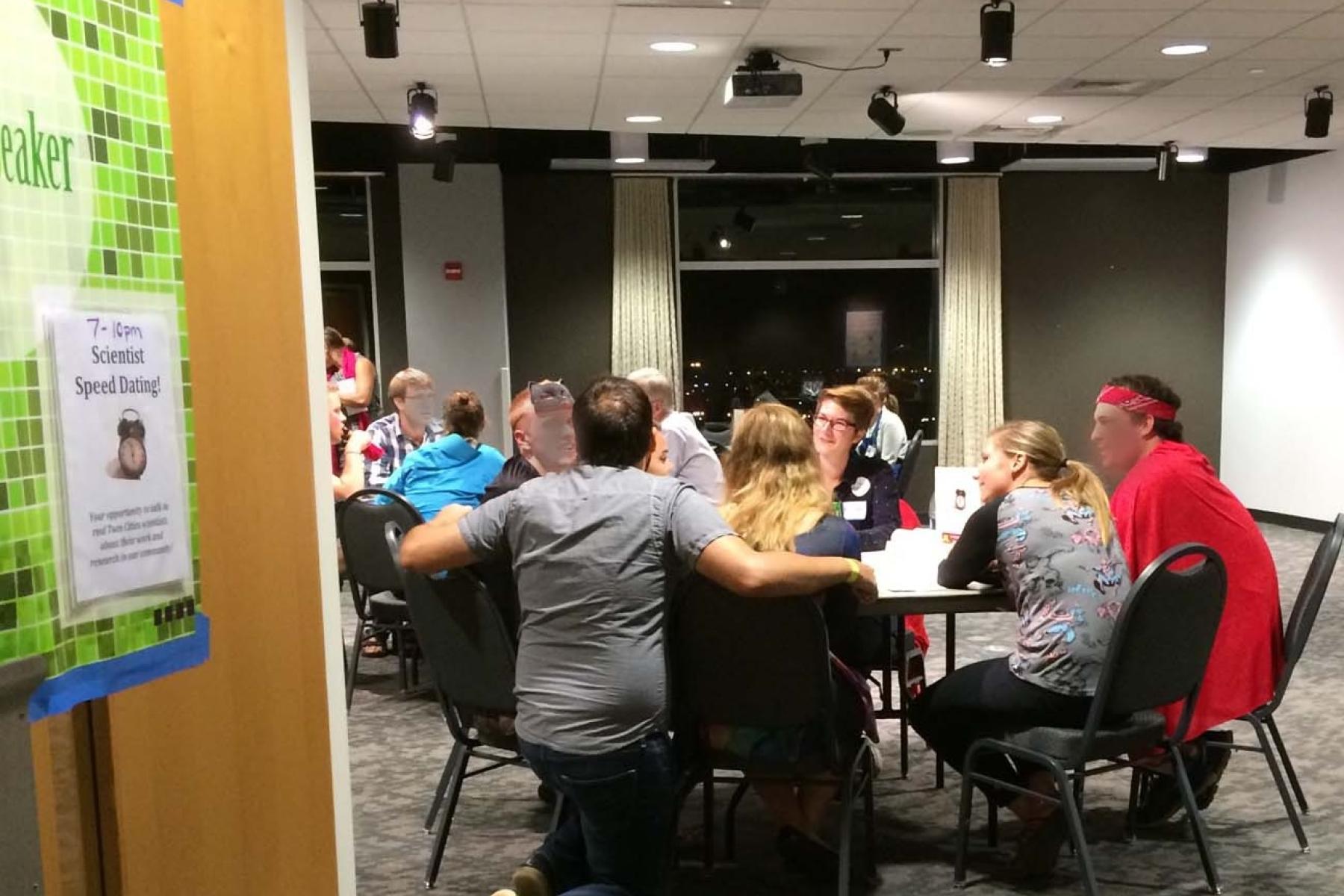 Peeking through the doorway to a large meeting room, there are multiple tables but the one closest to the door is most visible, there are about five or six people in a conversation. 