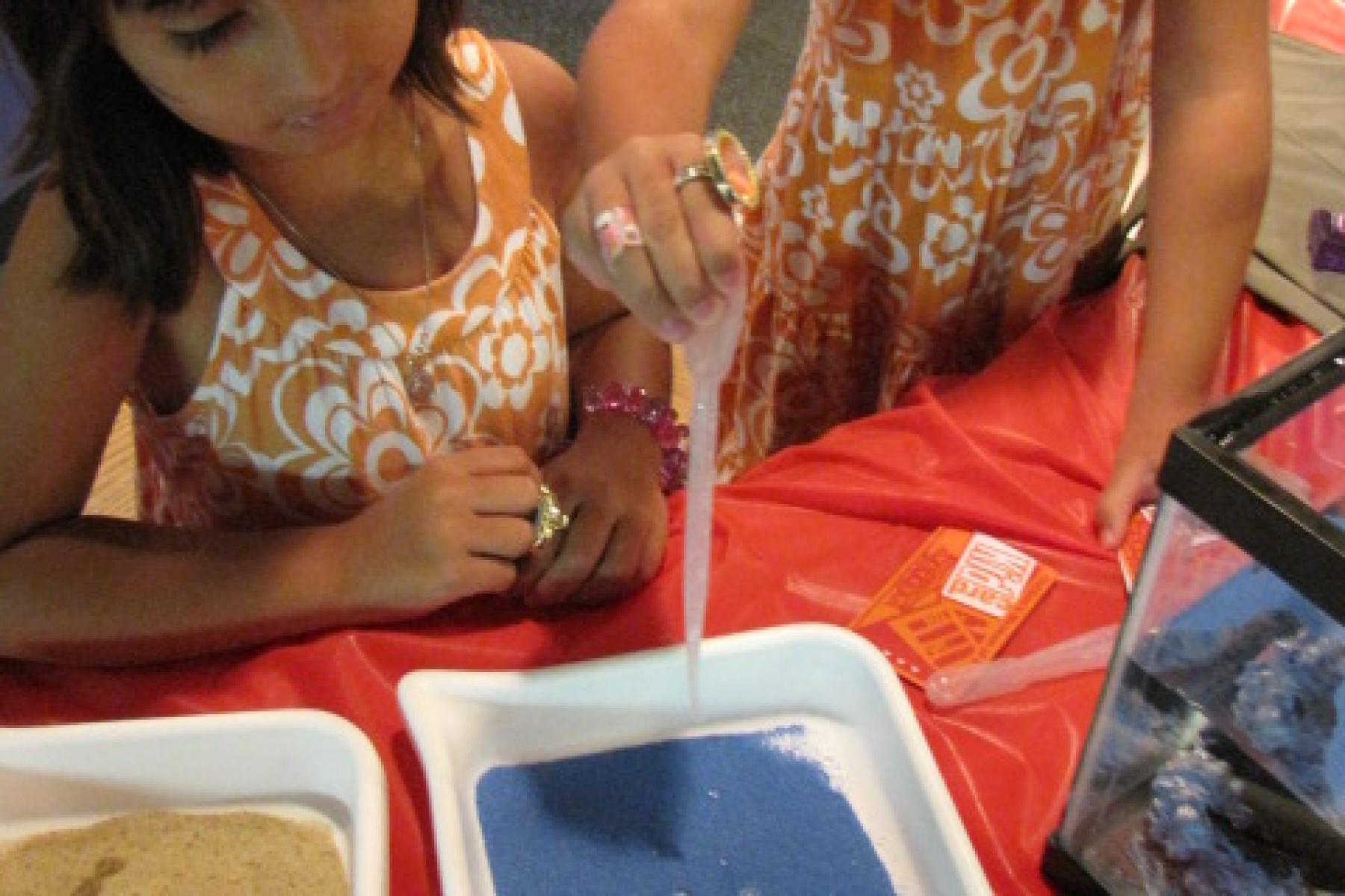 Two kids pouring water on blue hydrophobic sand in a white tray.